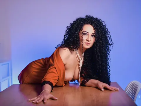 AnissiaTeasser — Glamorous woman with voluminous curly hair in a satin orange dress and pearls leaning on a wooden table.