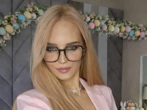 Blonde woman in pink blazer and glasses smiling in front of a floral background.