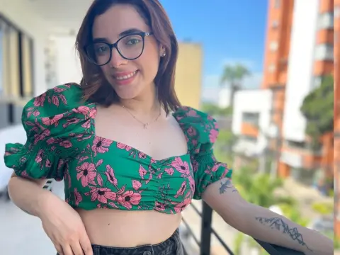 LiaCrimson smiles wearing a green floral puff-sleeve crop top and glasses, posing on a balcony with a city view.