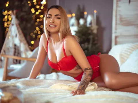 SamanthaRogue — Smiling blonde woman in red lace lingerie relaxing on a cozy bed in a warm, festive bedroom setting.
