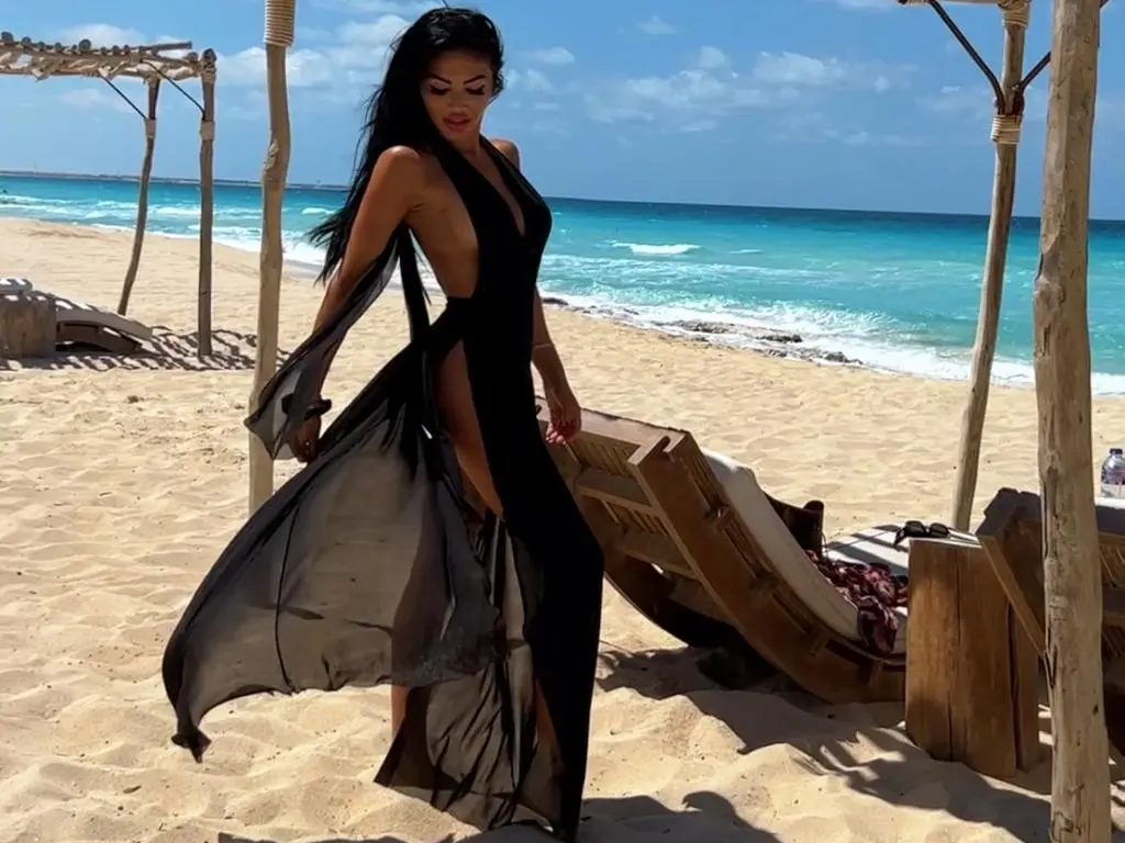 AlexxxaPower — Elegant woman in a flowing black maxi dress posing on a sunny tropical beach near wooden lounge chairs.