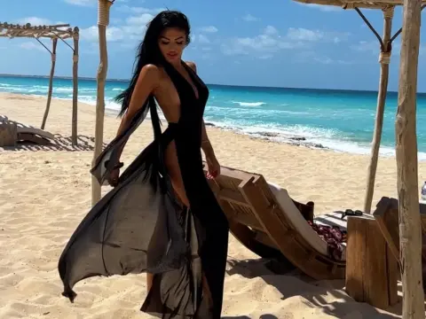 AlexxxaPower — Elegant woman in a flowing black backless maxi dress walking on a sunny tropical beach near wooden cabanas.