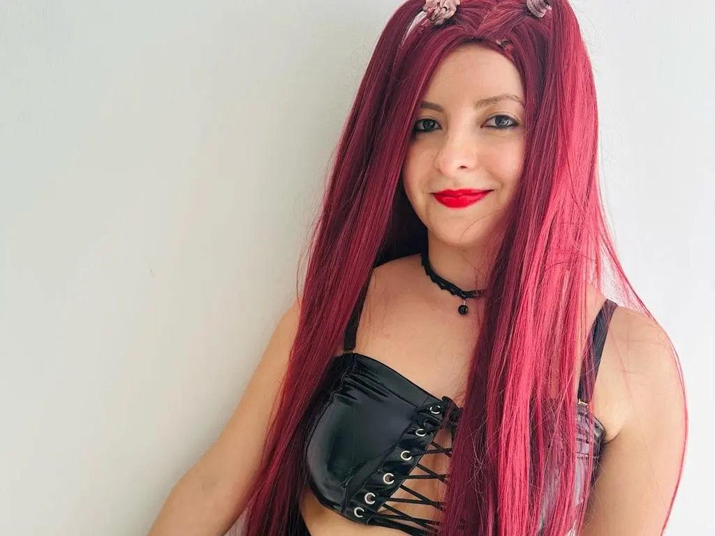 AlyssaFireston — AlyssaFireston smiles with long red hair and red lipstick, wearing a black lace-up corset and a black choker against a plain white wall.