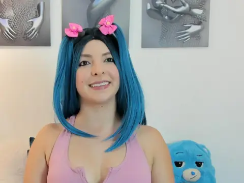 AlyssaFireston — AlyssaFireston smiles wearing a pink tank top and blue hair with pink flower clips, posing in front of grey wall art.