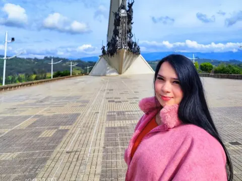 Smiling young woman in a pink fuzzy coat standing before a large monument under a blue sky.