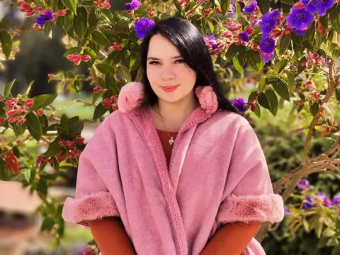 Smiling young woman in a pink fuzzy coat posing in front of vibrant purple flowers in a sunny garden.