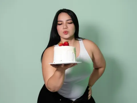 QueenSofia — QueenSofia wears a white tank top while holding a small white cake topped with strawberries against a green background.