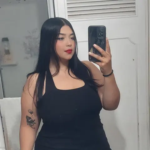 Young woman with long dark hair wearing a black tank top taking a mirror selfie in a bright room.