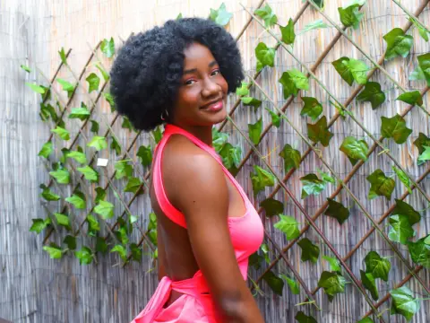 Smiling Black woman in a bright pink halter dress posing against a green ivy trellis background.