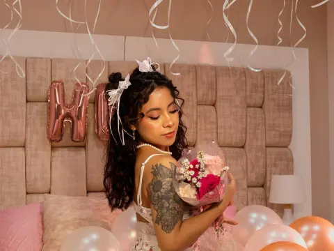 KarenMorgan — Young woman in white lace outfit holding pink flowers in a celebratory bedroom setting with balloons.