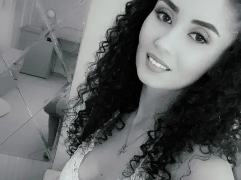 Smiling woman with long curly hair wearing a white lace top in a bright indoor setting.
