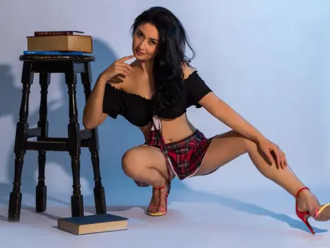 Young woman in black off-the-shoulder top and plaid skirt posing next to a wooden stool and books.
