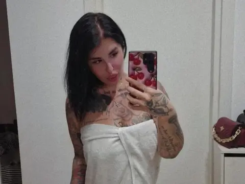 MallorieFoxx poses for a mirror selfie wearing a white tube top, showing off her dark hair and heavy arm and chest tattoos.