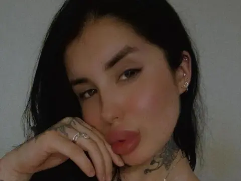 MallorieFoxx poses with her hand under her chin, showing her long dark hair, heavy eyebrows, and neck tattoos against a plain background.