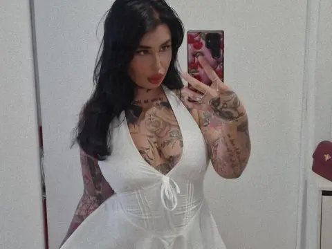 MallorieFoxx poses in a white corset top, showing off her long black hair and heavy tattoos on her chest and arms.