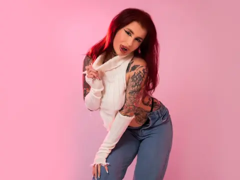 MallorieFoxx poses with her tongue out, wearing a white ribbed turtleneck sweater and blue jeans, showing off her tattoos and red hair against a pink background.