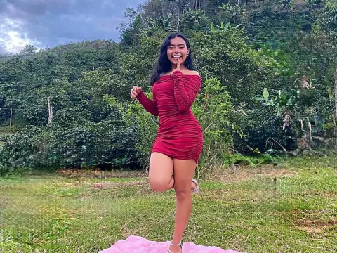 Julia_Bennett — Cheerful young woman in a red ruched mini dress posing playfully in a lush green forest setting.