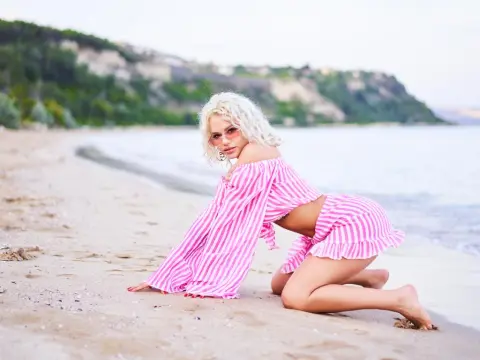 AstrydStorm poses on a sandy beach wearing a pink and white striped off-the-shoulder crop top and matching ruffled skirt, showing her blonde hair and sunglasses.