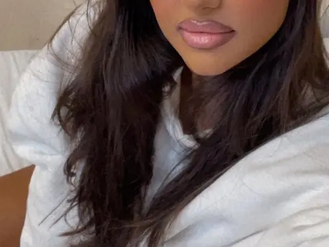 Dayana_Noir — Close-up of a woman with long dark hair and pink glossy lips wearing a casual white t-shirt in a bright setting.