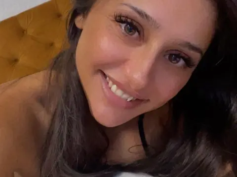 Dayana_Noir — Smiling young woman with long brown hair and natural makeup relaxing in a cozy indoor setting.