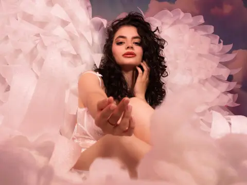 Dreamy woman in white lace lingerie with large glittery angel wings amidst soft pink clouds.