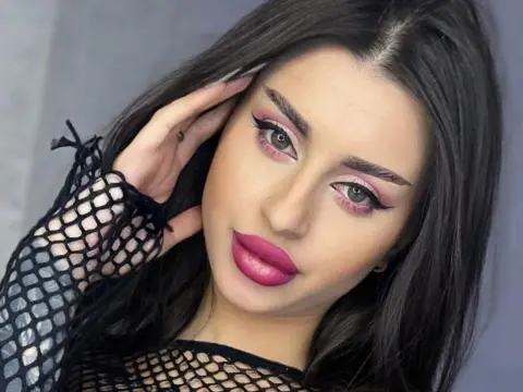 Young woman with long dark hair and pink makeup wearing a black fishnet top in a soft studio setting.