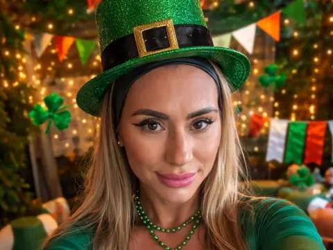 Smiling blonde woman in a green leprechaun hat and necklace celebrating St. Patrick's Day with festive lights.