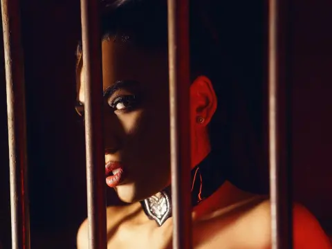 Moody portrait of a woman with dark makeup and a black choker looking through vertical bars in dramatic lighting.
