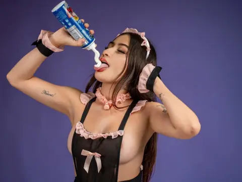 Playful woman in a pink ruffled black apron licking whipped cream from a blue canister against a purple background.