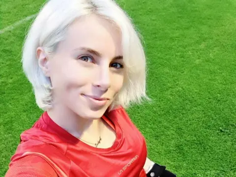 JennifferS — Smiling woman with short blonde hair wearing a red athletic shirt on a sunny green grass field.