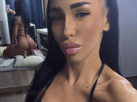 Young woman with long dark hair wearing black lingerie posing for a selfie in a bedroom setting.