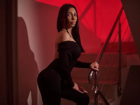 MyaBlakk — Elegant woman in a black long-sleeve outfit sitting on stairs under moody red lighting.