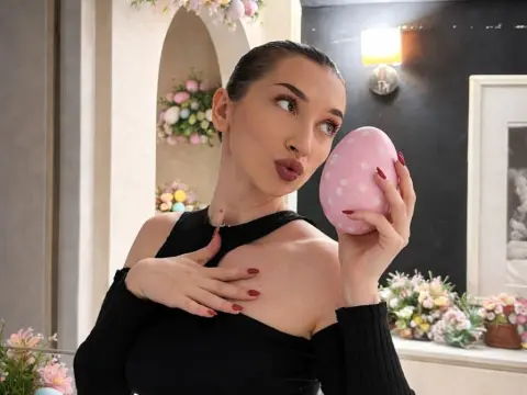 Elegant woman in a black off-the-shoulder top holding a pink polka dot Easter egg in a bright floral setting.