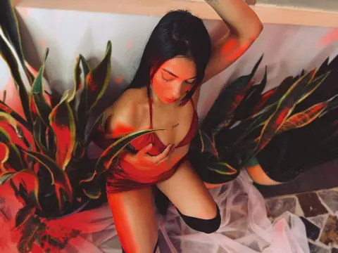 ashleesweett1 wears a red satin bodysuit while posing between large green leaves in a dimly lit setting.