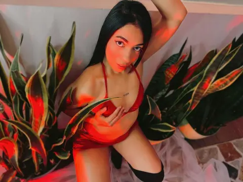 ashleesweett1 poses in a red satin bodysuit, surrounded by large green leaves and red lighting.