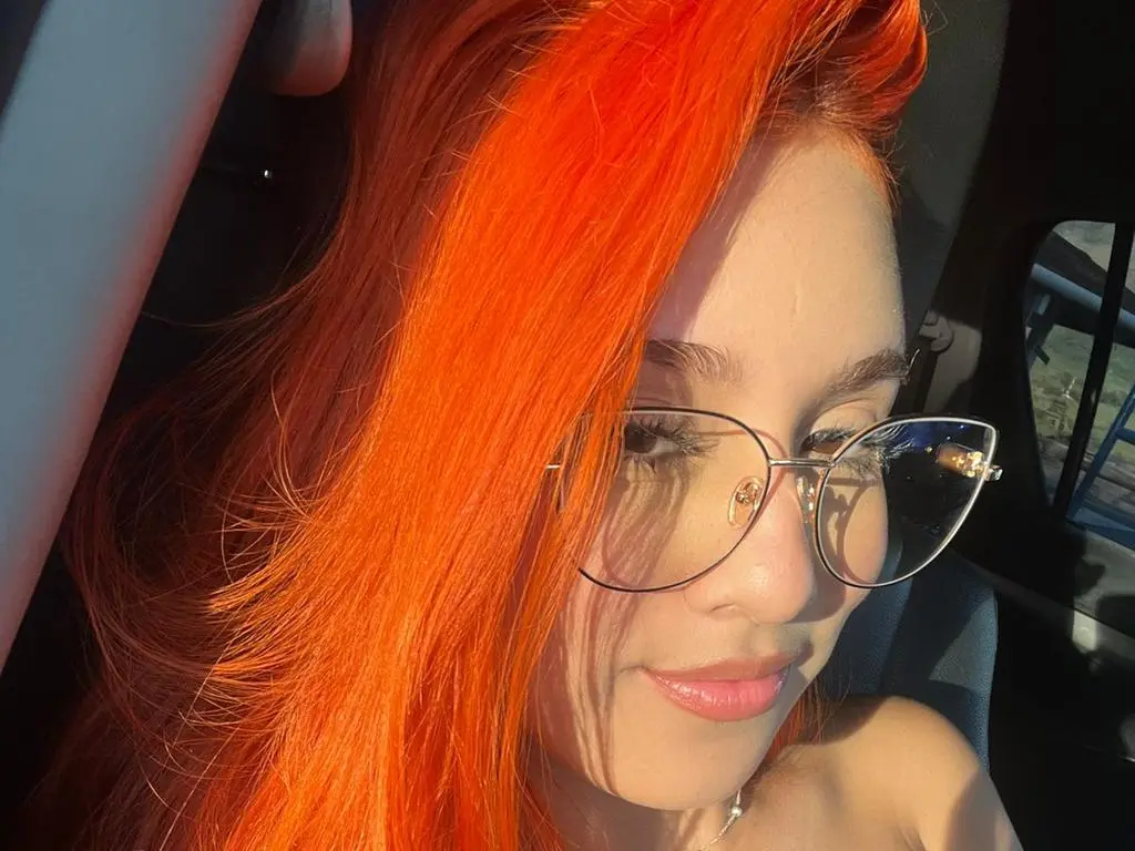 emmawatBuddy — Young woman with vibrant orange hair and wire-rimmed glasses smiling in a sunlit car interior.