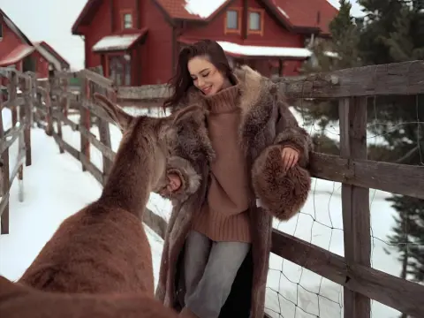 Smiling woman in a cozy brown sweater and fur coat petting a donkey in a snowy winter farm setting.