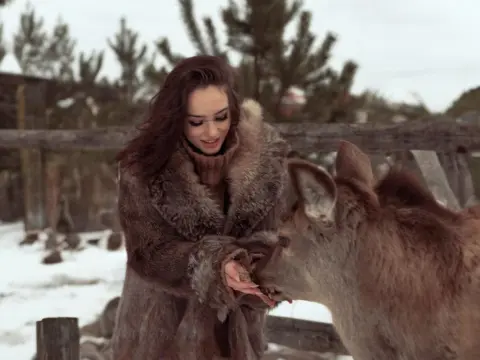 Smiling woman in a cozy brown faux fur coat feeding a deer in a snowy winter forest setting.