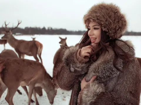 MissBelle — Smiling woman in a cozy brown fur coat and hat standing in a snowy field with deer in a winter landscape.