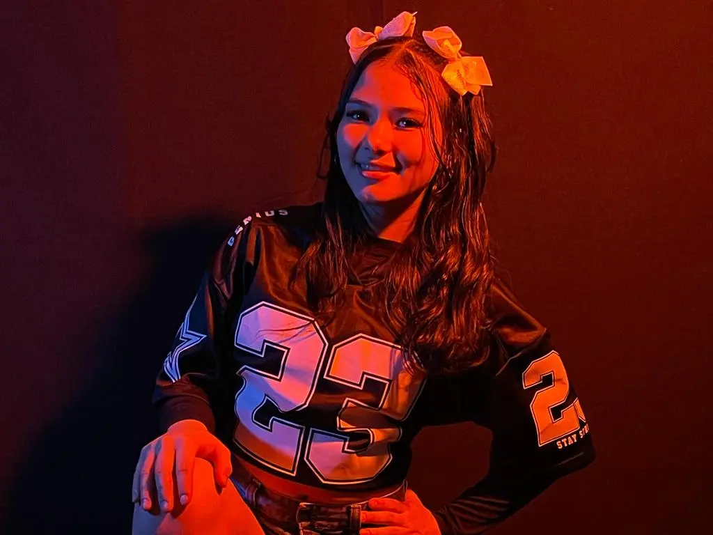 Smiling young woman in a black oversized jersey with white numbers posing under warm red studio lighting.