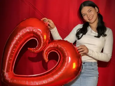 Smiling young woman in a white long-sleeve shirt holding a large red heart balloon against a red backdrop.