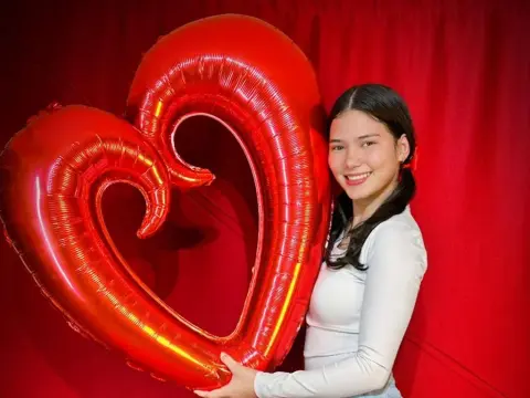 Smiling young woman in a white long-sleeve shirt holding a large red heart balloon against a red backdrop.