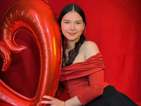 Smiling young woman in a red off-the-shoulder dress posing with a large red heart balloon against a red backdrop.