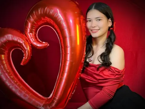 Smiling young woman in a red off-the-shoulder top posing with a large red heart balloon against a red backdrop.