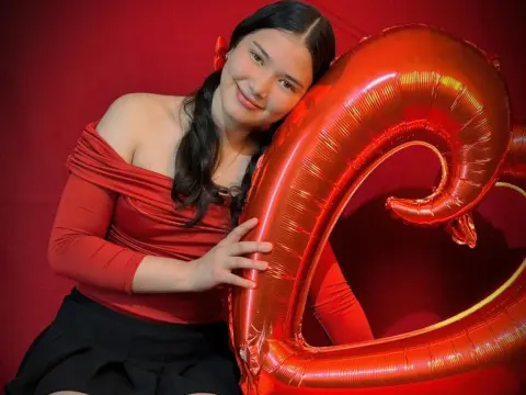 Smiling young woman in a red off-the-shoulder top leaning against a large red number 2 balloon against a red backdrop.