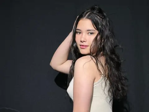 Young woman with long wavy hair wearing a white ribbed tank top against a dark studio background.