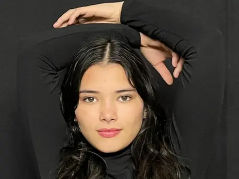 Young woman with long wavy hair wearing a black turtleneck against a dark background with a moody aesthetic.