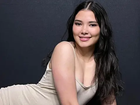 Smiling young woman with long dark hair wearing a beige ribbed tank top against a dark studio background.