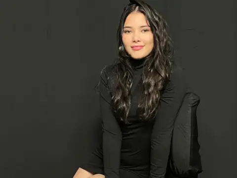 Smiling young woman in a black long-sleeve turtleneck dress posing against a dark studio background.