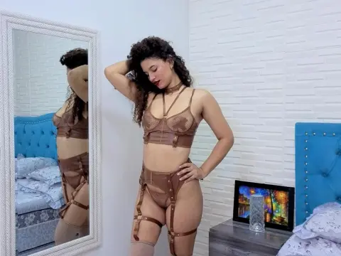 Curvy woman in a brown lace lingerie set posing in a bright bedroom with a white brick wall.